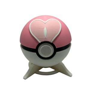 May include: A pink and white Poké Ball with a heart design on the top half. The ball has a black stripe and a gray button in the center. It is resting on a white stand with three legs.