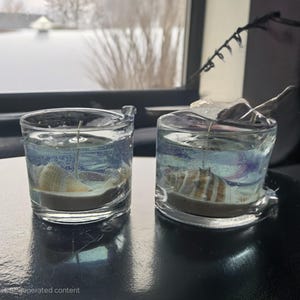 May include: Two clear glass candle jars with a seashell embedded in the center of each jar. The jars are filled with a clear gel and have a white wick.