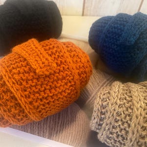 May include: Four hand-knitted beanies in various colors: black, orange, blue, and tan. The beanies are made of thick yarn and have a ribbed texture. The beanies are displayed in a white tray, with a neutral background.