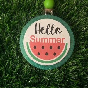 Puede incluir: Adorno redondo con temática de verano con la inscripción "Hello Summer". Presenta un diseño de sandía con una rodaja roja, semillas negras y corteza verde. El adorno está suspendido por una cuerda de yute y una cuenta verde, de unos 7 cm de diámetro.