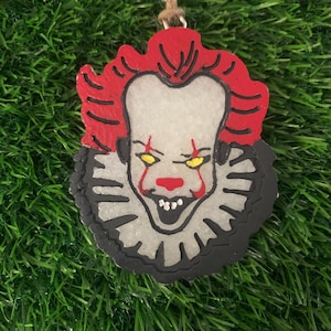 May include: A decorative ornament featuring a stylized image of a clown's face with red hair, a white face, and a black and white ruffled collar. The clown has yellow eyes and a red nose and mouth. The ornament is hanging from a brown string.