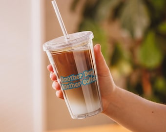 May include: A clear plastic tumbler with a lid and straw, filled with iced coffee. The tumbler has the text "Another Day Another Coffee" printed on it in blue and yellow.