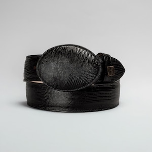 May include: A black leather belt with a textured finish. The belt features a large, oval buckle and a matching belt loop. The buckle has a reptile-like pattern. The belt is displayed against a plain white background.