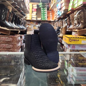 May include: Black leather ankle boots with a textured, scale-like pattern. The boots have a square toe and a low heel with a light-colored sole. The image shows the boots in a store setting, with other boots visible in the background.