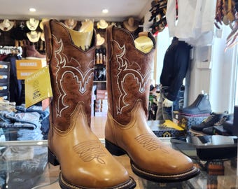 May include: A pair of tan leather cowboy boots with brown suede tops. The boots have intricate white stitching and a pointed toe. The boots are on a display shelf in a store.