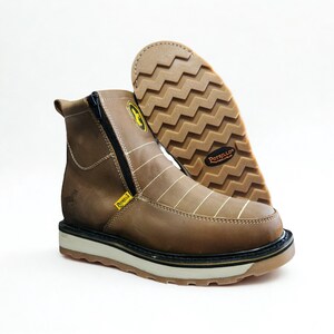 May include: Brown leather work boot with a side zipper and white stitching. The boot has a thick, textured sole and a yellow logo. The sole of the other boot is visible, showing a zig-zag pattern.