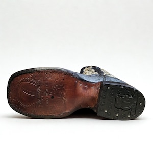 May include: A close-up of the sole of a cowboy boot, revealing the craftsmanship. The boot has a dark brown leather sole with embossed patterns and a black rubber heel. The upper part of the boot is visible, with a patterned design.