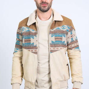 May include: A beige bomber jacket with a brown suede yoke and a cream-colored shearling collar. The jacket features a Southwestern-inspired pattern in shades of blue, brown, and cream across the chest. The jacket has a zipper closure and ribbed cuffs.