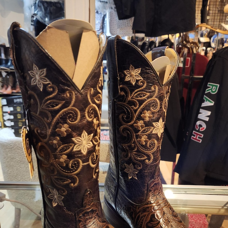 May include: A pair of dark brown cowboy boots with detailed floral embroidery in gold and cream thread. The boots have a pointed toe and a tall shaft, with a textured leather appearance. The boots are displayed on a glass surface.