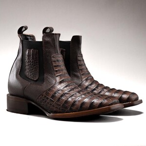 May include: A pair of brown leather Chelsea boots with a crocodile skin pattern. The boots have a low heel and elastic side panels. The pull tabs are visible at the top of the boots. The boots are made of a textured leather material.
