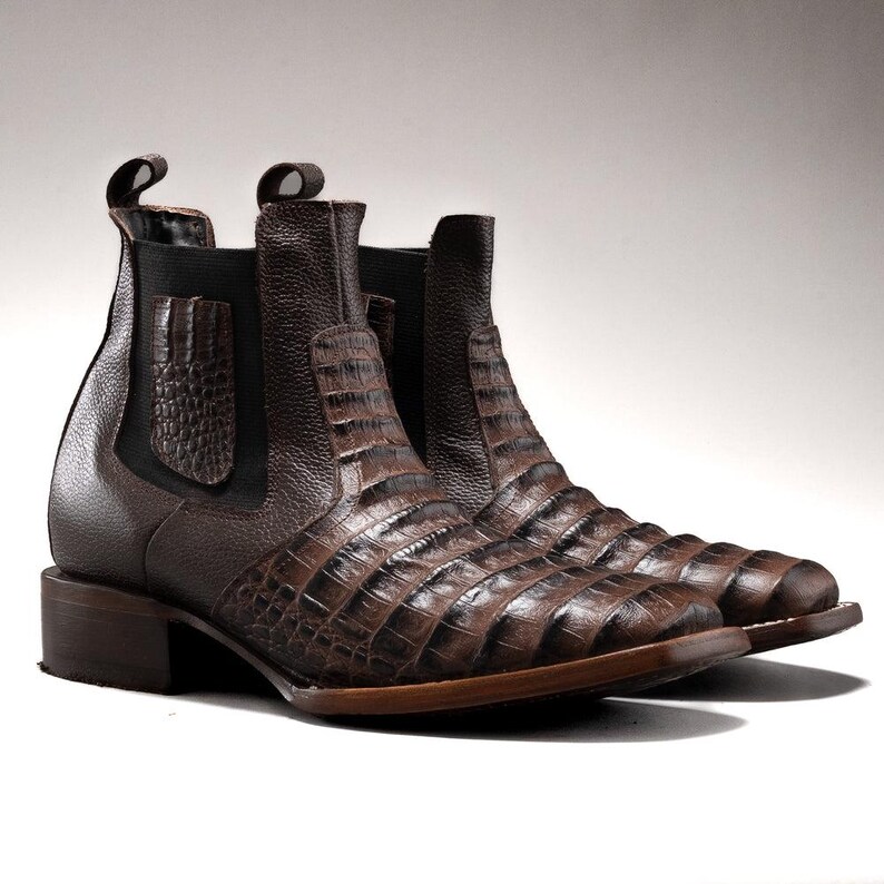 May include: A pair of brown leather Chelsea boots with a crocodile skin pattern. The boots have a low heel and elastic side panels. The pull tabs are visible at the top of the boots. The boots are made of a textured leather material.