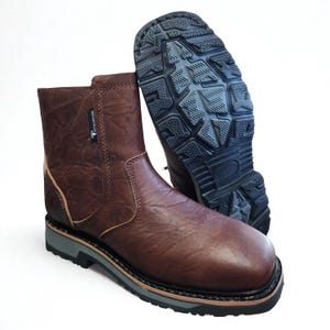 May include: A brown ankle boot with a textured surface and a small tag labeled waterproof. The boot features a thick black lugged outsole with a grey midsole layer. A second boot is positioned behind it, showing the black tread pattern on the sole.