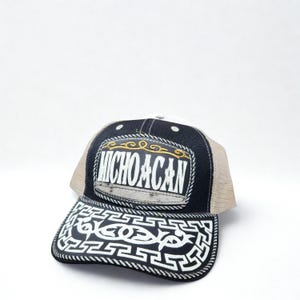 May include: A black and tan trucker hat with the word "MICHOACAN" embroidered in white. The hat features a decorative design on the brim and a white mesh back.