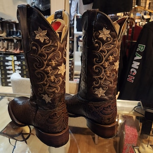 May include: Two brown leather cowboy boots with detailed floral embroidery and decorative patterns. The boots feature a pointed toe and a stacked heel, reflecting a classic Western style. The boots are displayed on a glass surface.