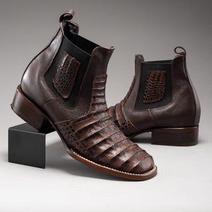 May include: A pair of brown leather ankle boots with a crocodile skin pattern. The boots have a square toe, elastic side panels, and a pull tab at the back. The heels are approximately 1.5 inches high.