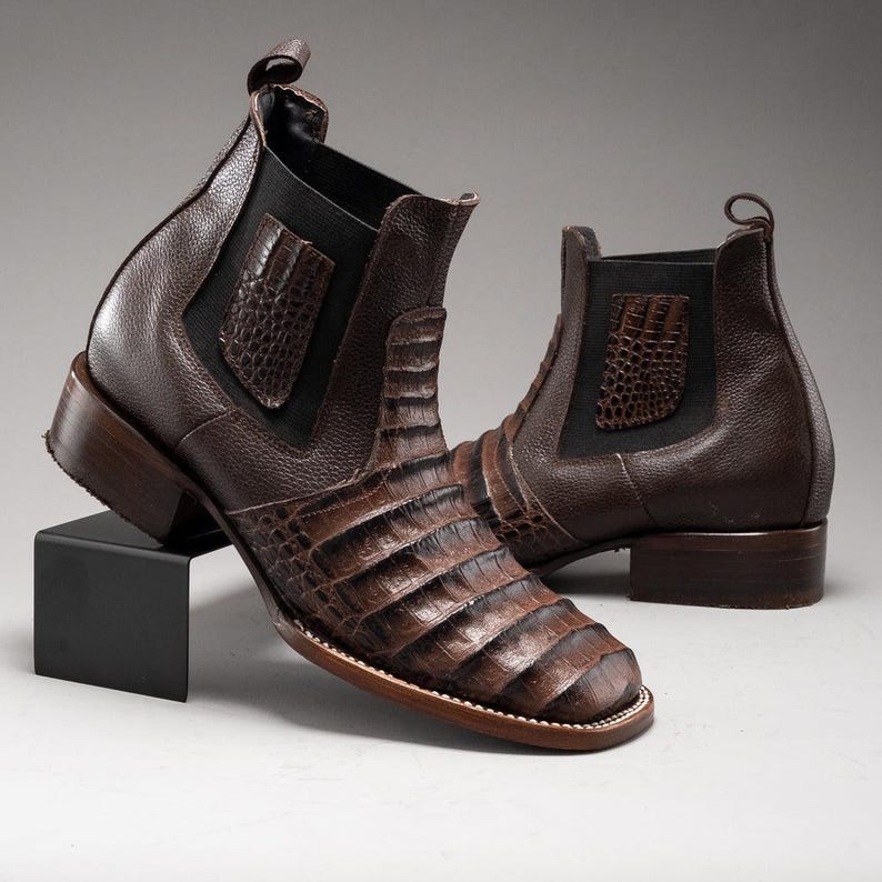 May include: A pair of brown leather ankle boots with a crocodile skin pattern. The boots have a square toe, elastic side panels, and a pull tab at the back. The heels are approximately 1.5 inches high.