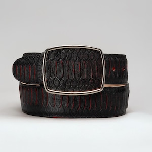 May include: Black belt with a textured pattern and red accents. The belt features a silver-colored rectangular buckle. The belt is made of a material that resembles snakeskin.
