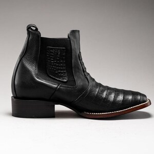 May include: Black leather ankle boot with a square toe and a low block heel. The boot features a textured pattern on the vamp and a pull tab at the back. The sole has a brown edge with white stitching.