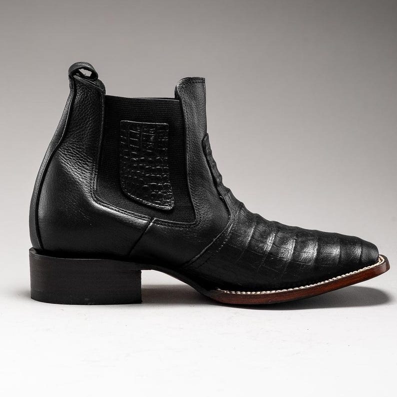 May include: Black leather ankle boot with a square toe and a low block heel. The boot features a textured pattern on the vamp and a pull tab at the back. The sole has a brown edge with white stitching.