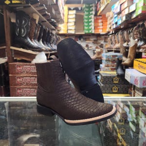 May include: Brown leather ankle boot with a textured, scale-like pattern. The boot has a square toe and a black sole. The image shows the sole of a second boot. The background shows a shoe store with various cowboy boots.