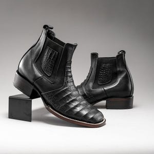 May include: A pair of black leather ankle boots with a square toe and a low heel. The boots feature a crocodile-textured design on the vamp and a smooth leather upper. The boots are displayed on a black stand against a neutral background.