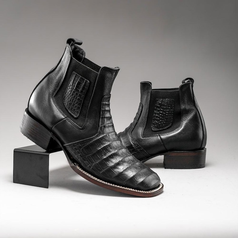 May include: A pair of black leather ankle boots with a square toe and a low heel. The boots feature a crocodile-textured design on the vamp and a smooth leather upper. The boots are displayed on a black stand against a neutral background.