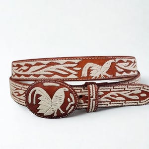May include: A brown leather belt with a decorative buckle featuring a rooster design. The belt has white embroidered patterns and stitching. The buckle is oval-shaped and the belt is approximately 1.5 inches wide.