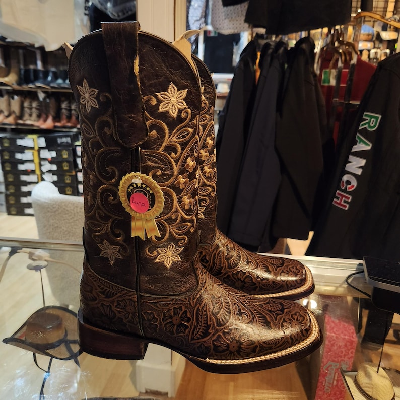 May include: A pair of brown leather cowboy boots with detailed floral and scroll embroidery. The boots have a square toe, a stacked heel, and a decorative tag. The boots are displayed on a glass surface.