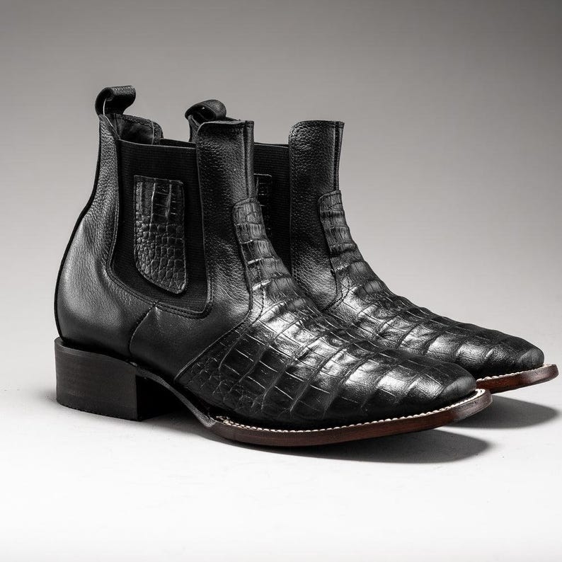 May include: A pair of black leather ankle boots with a crocodile skin pattern. The boots have a square toe, a low heel, and elastic side panels. The boots are approximately 6 inches tall.