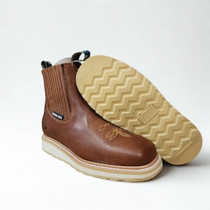 May include: Brown leather Chelsea boots with a textured upper and a white sole. The boots feature a decorative stitch design on the front and a pull tab with green accents. The sole is a light tan color with a zig-zag pattern.