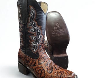 May include: Brown and black cowboy boots with floral and scrollwork designs. The boots have a square toe and a stacked heel. The shaft is black with embroidered details. The sole of the boot is visible, with the brand name "Potrillo" stamped on it.