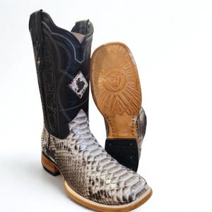 May include: A pair of cowboy boots with a snakeskin pattern on the lower part and a black leather upper. The boots have a square toe and a decorative diamond-shaped emblem. The sole of one boot is visible, showing a detailed design.