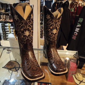 May include: A pair of brown leather cowboy boots with detailed floral and scrollwork embroidery. The boots feature a square toe, a decorative buckle, and a light-coloured sole. The boot shafts are tall, and the overall design is rustic and detailed.