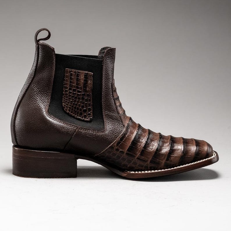 May include: A brown leather ankle boot with a crocodile-textured vamp and a pull tab. The boot features a square toe and a stacked heel. The side panel is elasticized for easy wear. The boot's overall design is sleek and stylish.