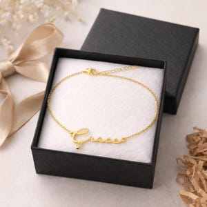 Puede incluir: Un brazalete dorado con el nombre "Grace" en escritura cursiva, presentado en una caja de regalo negra abierta con interior blanco. El brazalete muestra una fina cadena y cierre. El fondo incluye una cinta dorada y elementos decorativos.