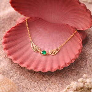 May include: A gold-colored necklace with a wing design and a green gemstone center. The necklace is displayed on a pink seashell, set against a sandy background with other seashells and small white flowers.