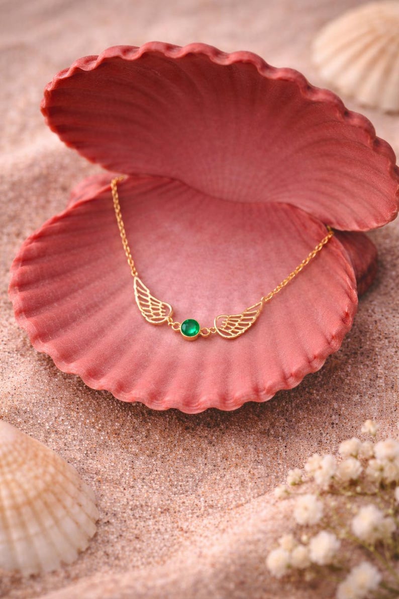 May include: A gold-colored necklace with a wing design and a green gemstone center. The necklace is displayed on a pink seashell, set against a sandy background with other seashells and small white flowers.