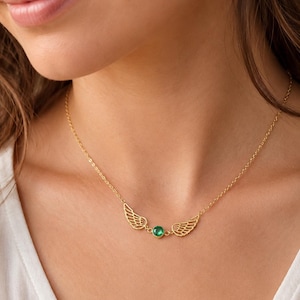 May include: A gold-colored necklace featuring a pair of openwork wings on either side of a round, green gemstone. The necklace is worn around the neck, with the chain visible. The background is a soft, neutral tone.