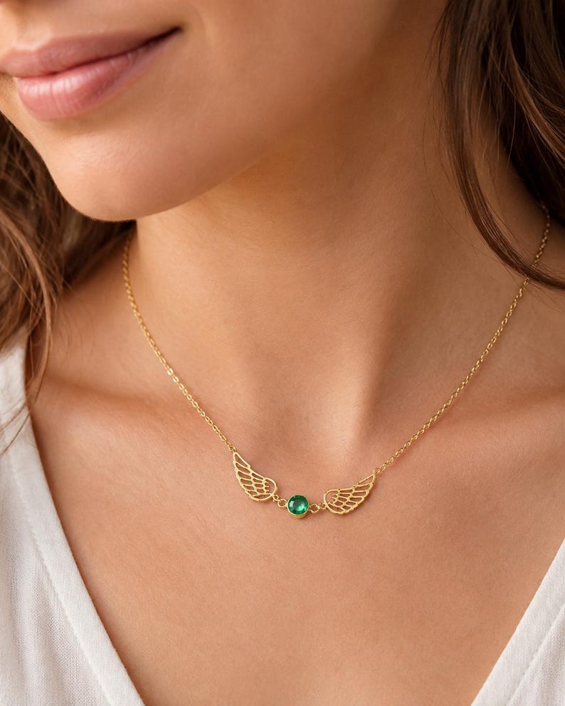 May include: A gold-colored necklace featuring a pair of openwork wings on either side of a round, green gemstone. The necklace is worn around the neck, with the chain visible. The background is a soft, neutral tone.