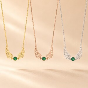 May include: Three necklaces with wing pendants and a green gemstone. The necklaces are gold, rose gold, and silver. The pendants have a detailed wing design and a round, green gemstone in the center.