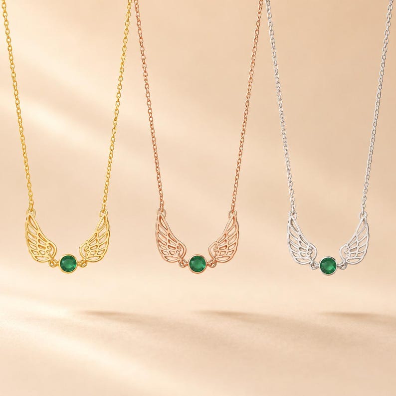May include: Three necklaces with wing pendants and a green gemstone. The necklaces are gold, rose gold, and silver. The pendants have a detailed wing design and a round, green gemstone in the center.