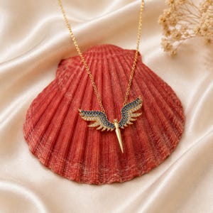 May include: A gold-colored necklace with a pendant shaped like angel wings. The wings are detailed with blue and clear gemstones. The necklace is displayed on a red seashell against a cream-colored satin background.