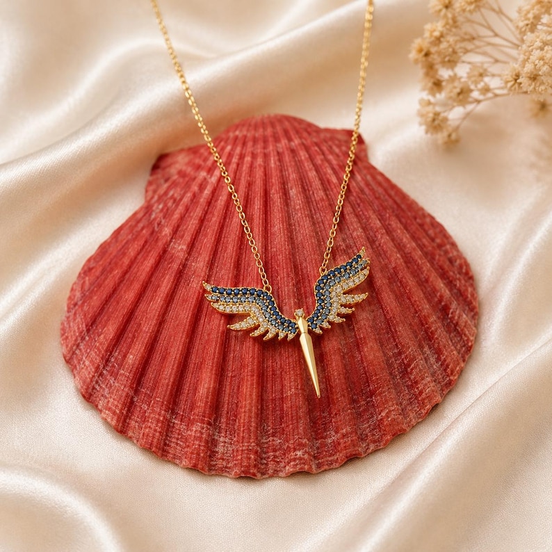 May include: A gold-colored necklace with a pendant shaped like angel wings. The wings are detailed with blue and clear gemstones. The necklace is displayed on a red seashell against a cream-colored satin background.