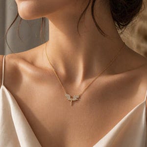 May include: A delicate gold necklace featuring a pendant with an angel wing design. The wings are adorned with small, sparkling stones. The necklace is displayed on a person wearing a cream-colored top.