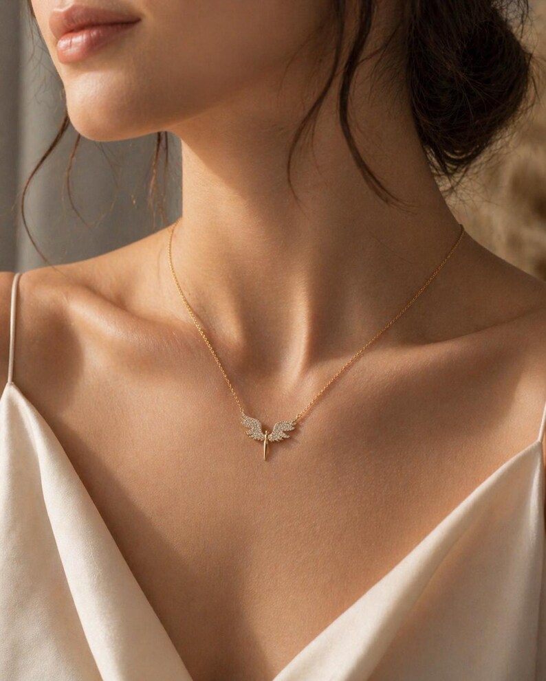 May include: A delicate gold necklace featuring a pendant with an angel wing design. The wings are adorned with small, sparkling stones. The necklace is displayed on a person wearing a cream-colored top.