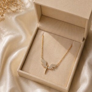 May include: A gold-colored necklace with an angel wing pendant, displayed in a beige jewelry box. The pendant features a central spike and two wings encrusted with small, sparkling stones. The necklace chain is delicate and gold-toned.