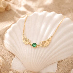 May include: A gold-colored necklace with a pair of wing-shaped pendants and a central, round, green gemstone. The necklace is displayed on a white seashell, set against a sandy background.
