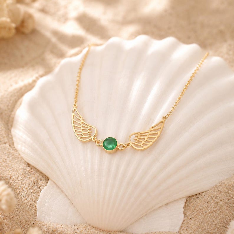 May include: A gold-colored necklace with a pair of wing-shaped pendants and a central, round, green gemstone. The necklace is displayed on a white seashell, set against a sandy background.