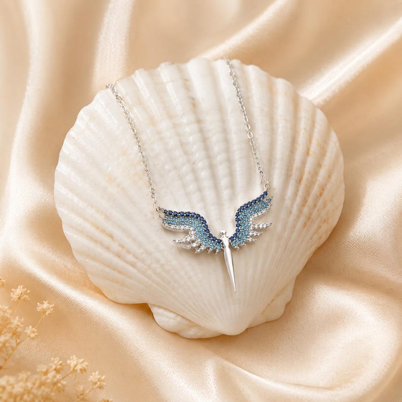 May include: A silver necklace with a pendant shaped like angel wings, adorned with blue and clear gemstones. The necklace is displayed on a white seashell against a backdrop of a satin fabric.