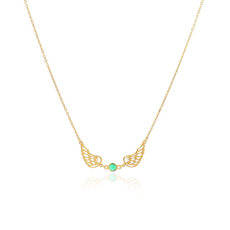 May include: A gold-colored necklace featuring a pair of openwork wings on either side of a small, round, green gemstone. The necklace chain is delicate and thin, and the wings have intricate detailing.